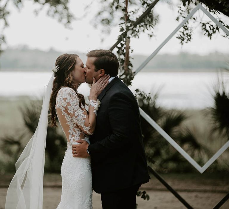 Bride and groom share their first kiss as a married couple with bride in floral lace illusion wedding dress with sleeves and groom in black suit jacket