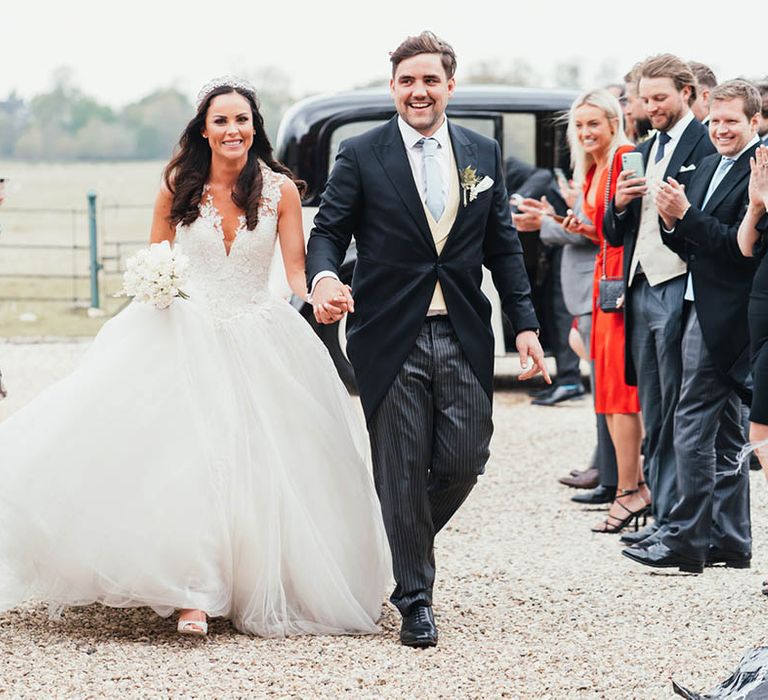 Bride in princess wedding dress with white rose bouquet walks inbetween guests with groom in morning suit with pale blue tie 