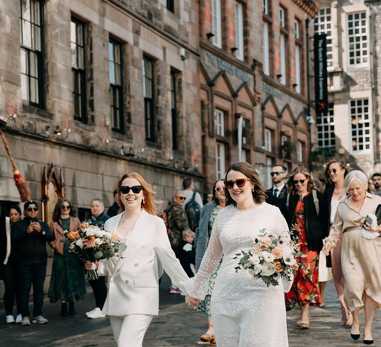 Brides wearing sunglasses walk with one another on their wedding day holding hands and carrying floral bouquets 