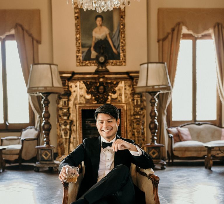 Groom wears black tie and sits in rustic chair whilst holding glass of whiskey on the morning of his wedding day