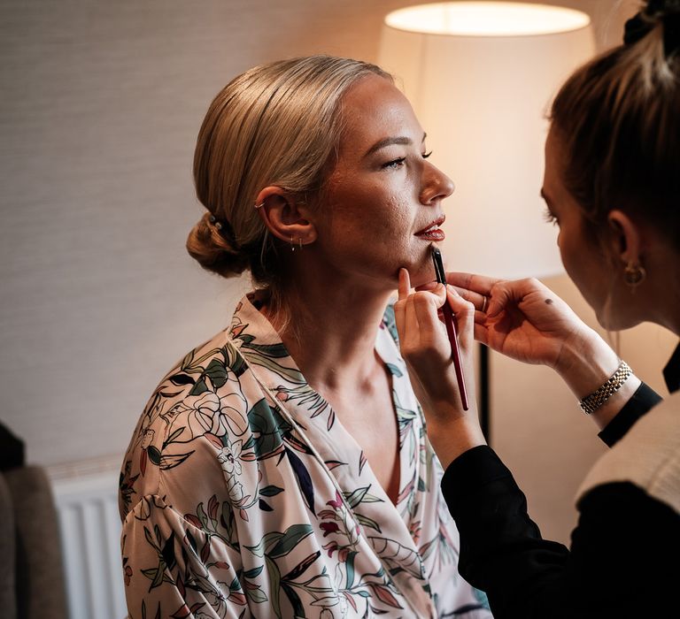 Bride wears her blonde hair slicked back into low bun complete with pearl hair accessories as makeup artist applies lipstick to her