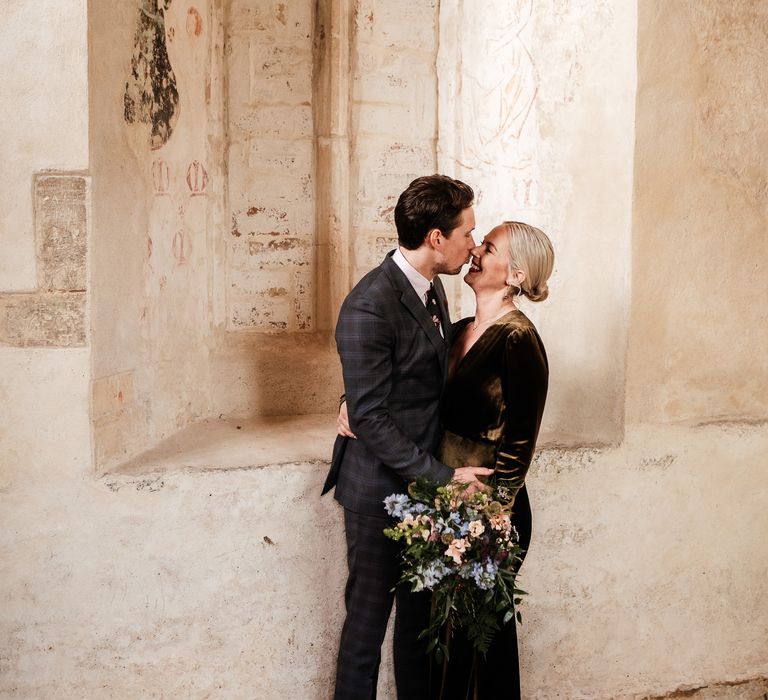 Bride wears her blonde hair slicked into low bun complete with pearl hair accessories as she kisses her groom who wears tartan suit 