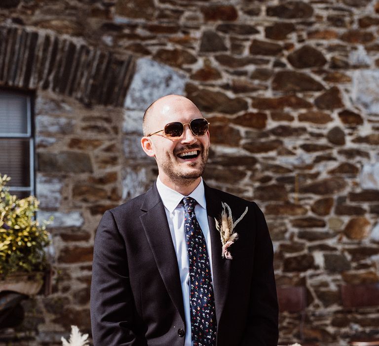 Groom smiles wearing brown sunglasses in a navy blue suit and an easter bunny tie 