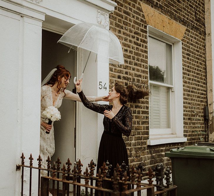 Bride leaves flat whilst bridesmaid wearing black bridesmaid lace dress carries see through umbrella above her head