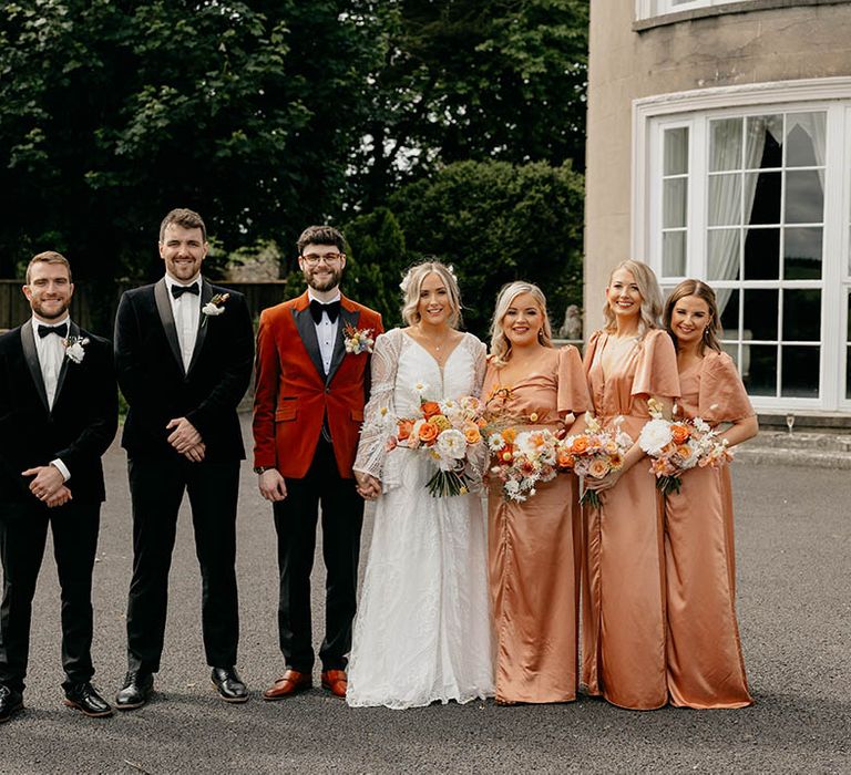 Wedding party shot with groomsmen in black tie, bridesmaids in peach satin dresses and bride and groom 