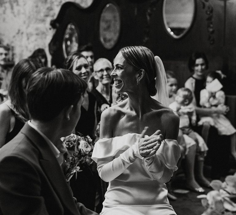 Bride in an off the shoulder satin wedding dress smiles at the guests as she sits with the groom during their wedding ceremony 