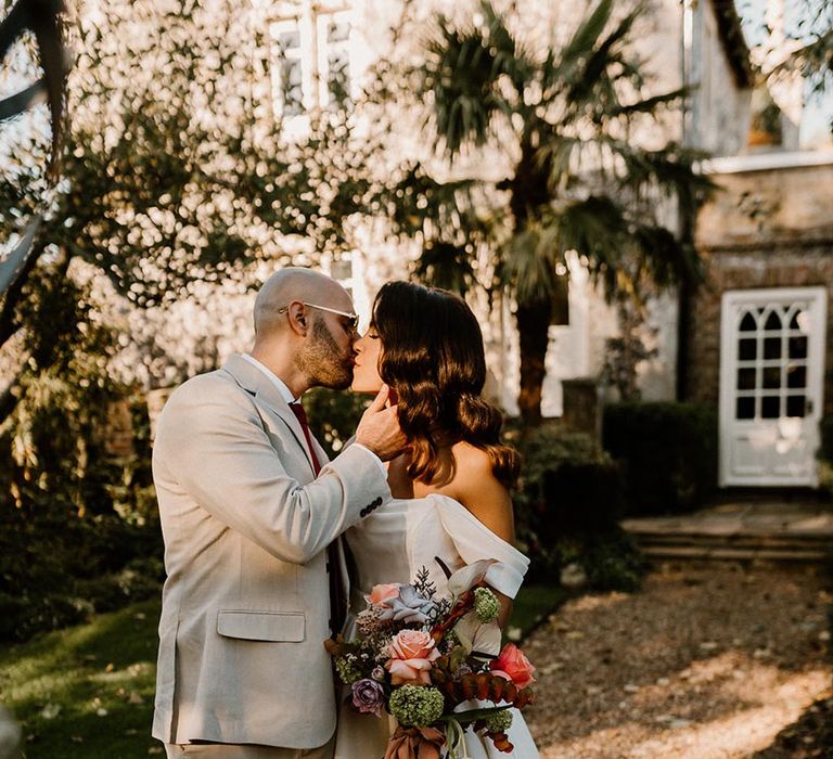 Groom in a pale grey su8it and dark red tie kisses the bride in an off the shoulder wedding dress and Hollywood waves