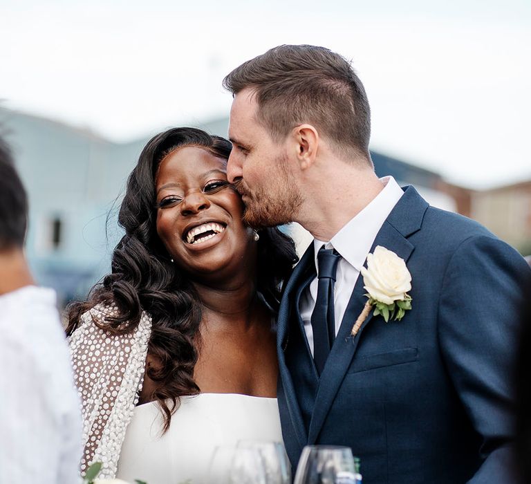 Bride wears her black hair in loose curls with Suzanne Neville wedding dress as her groom in three-piece suit kisses her cheek