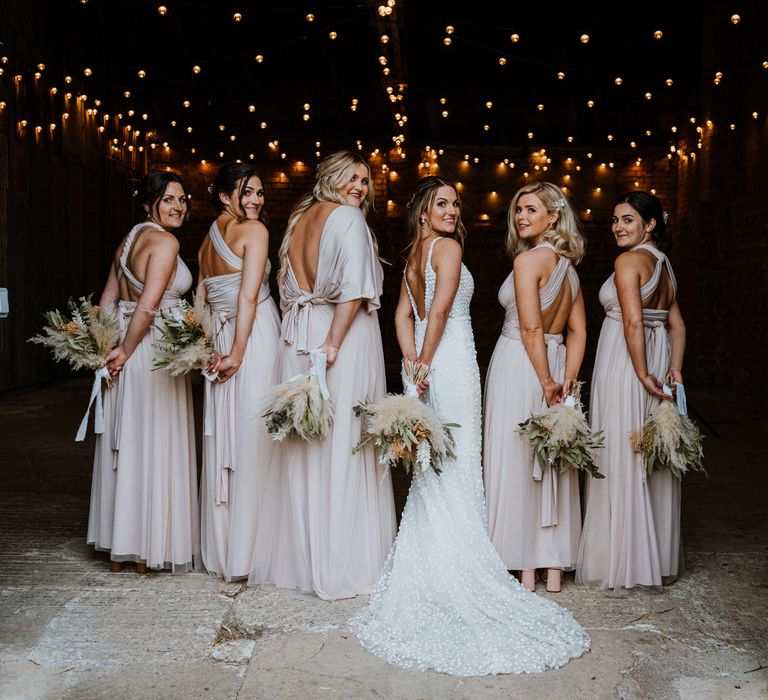 Bridal party turning back to the camera wearing light cream bridesmaid dresses with open backs and bride wearing sleeveless Made With Love Bridal dress with plunging back all holding dried flower bouquets 