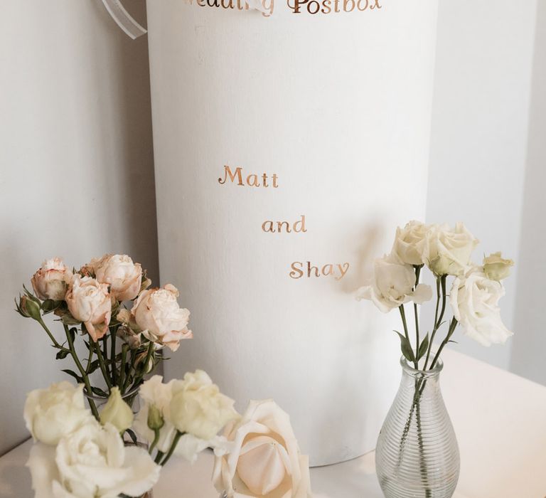 White & gold painted wedding postbox finished with white ribbon surrounded by florals