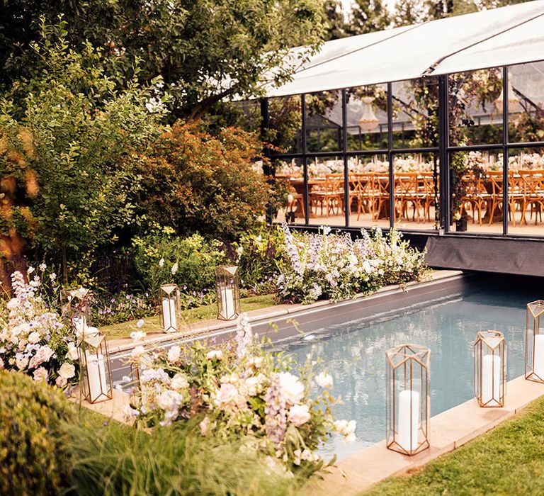 Pool area with candles in lanterns, pink and white flowers, and glass marquee reception area for at home wedding 