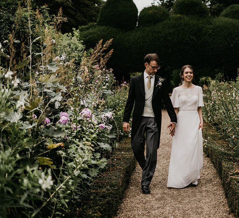 Bride in batwing sleeve lace detail wedding dress and groom in three piece suit with light waistcoat walking around the grounds of Kelmarsh Hall country house