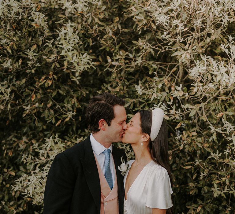 Groom in morning suit with colourful waistcoat and tie kissing the bride in a satin button wedding dress and chunky pearl headband