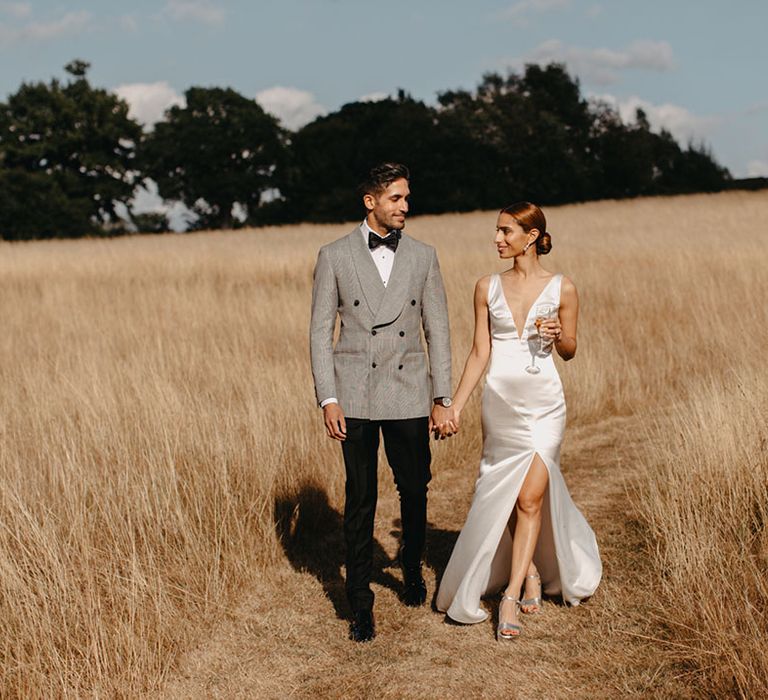 Bride in silk wedding dress walks through golden fields with her groom in black-tie on her wedding dress