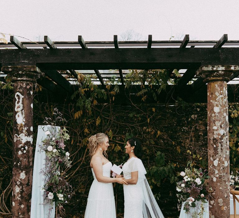 Brides in sleeveless lace wedding dress and off the shoulder long sleeve wedding dress holding hands at the alter with mixed floral decorations with white carnations, purple tulips, roses and foliage 