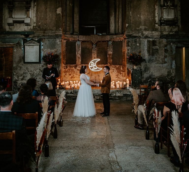 Bride and Groom stand at alter with moon neon sign in the centre at industrial wedding venue in London