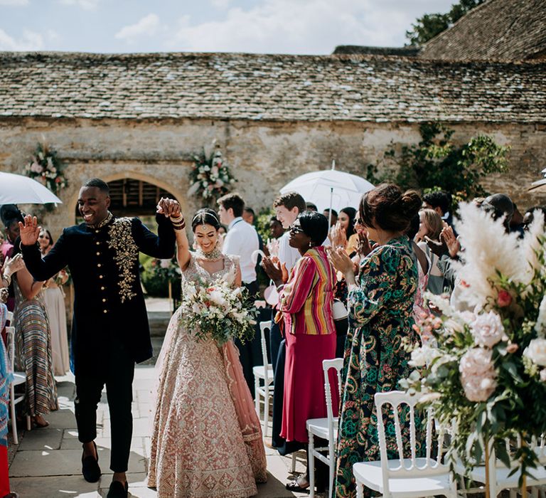 Panjabi Bride and Jamaican Groom walk up the aisle holding hands in golden bridal Lehnga and midnight blue and gold Indian groom outfit