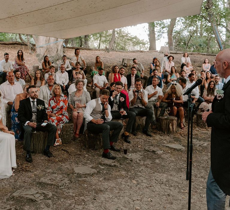 Groomsman reads out a wedding speech as the bride and groom and wedding guests sit and listen in secret outdoor area 