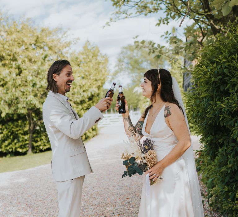 Bride and groom in ASOS wedding outfits and black high-top converse cheers-ing their beer bottles