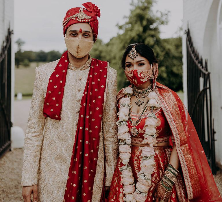 Bride & groom in matching red and gold sherwani and lehenga wear masks outdoors on their wedding day