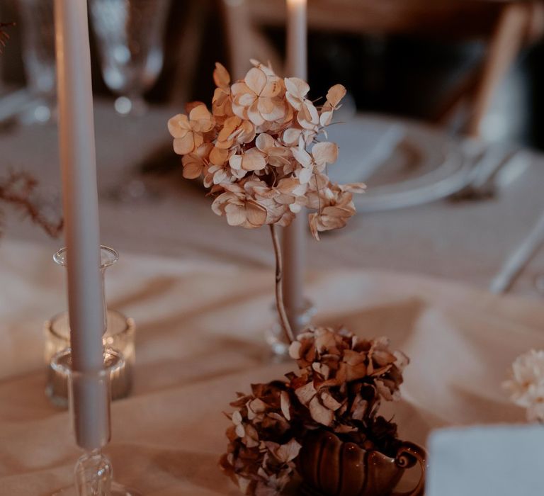 Beige tapered candles in clear candlestick holders with dried light pink and blush dried flower arrangements and tea light candles in textured beige candleholders on minimalistic white tablecloth 