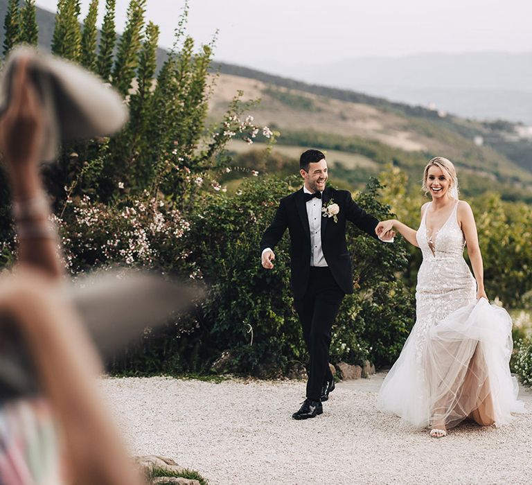 Bride in lace wedding dress and trumpet styled ruffled skirt walks alongside her groom in black-tie