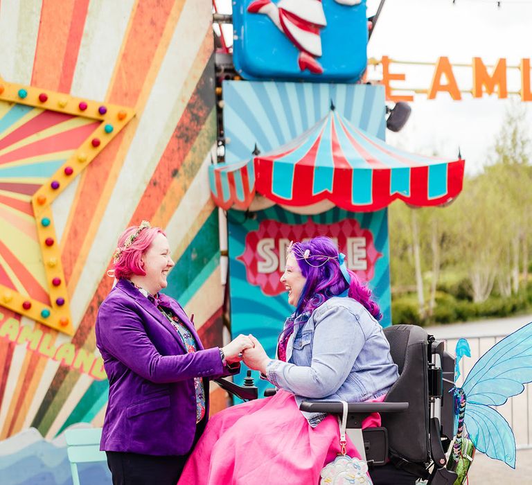 Bride in wheelchair with sparkly blue fairy wings attached wearing personalised denim jacket, pink wedding dress and blue tights holding hands with bride in  purple velvet blazer and black suit trousers with purple bowtie, pink patterned pocket square and colourful patterned shirt 