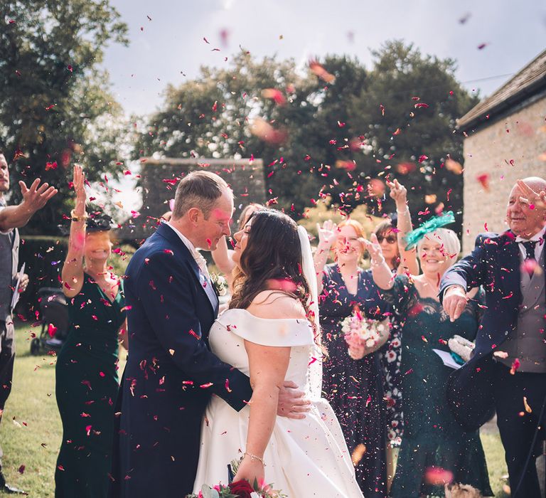 Groom in navy morning suit leaning in for a kiss with the bride in an off the shoulder wedding dress holding a blue and purple bouquet with peony confetti 