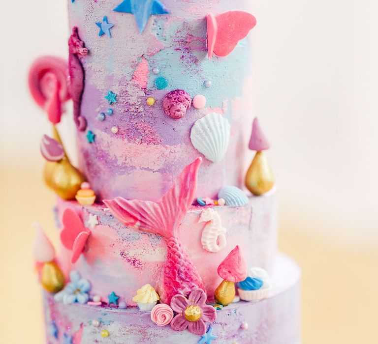 Four tier pink and purple wedding cake with pink mermaid tail, lollipops, stars, butterflies, shells and other small icing details 