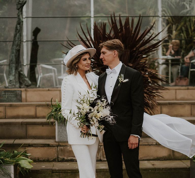 Bride wears Nadine Merabi bridal suit with pearl embellishment and white bridal hat complete with white floral bouquet 