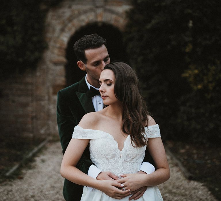 Bride in off the shoulder lace wedding dress being embraced from behind by the groom 