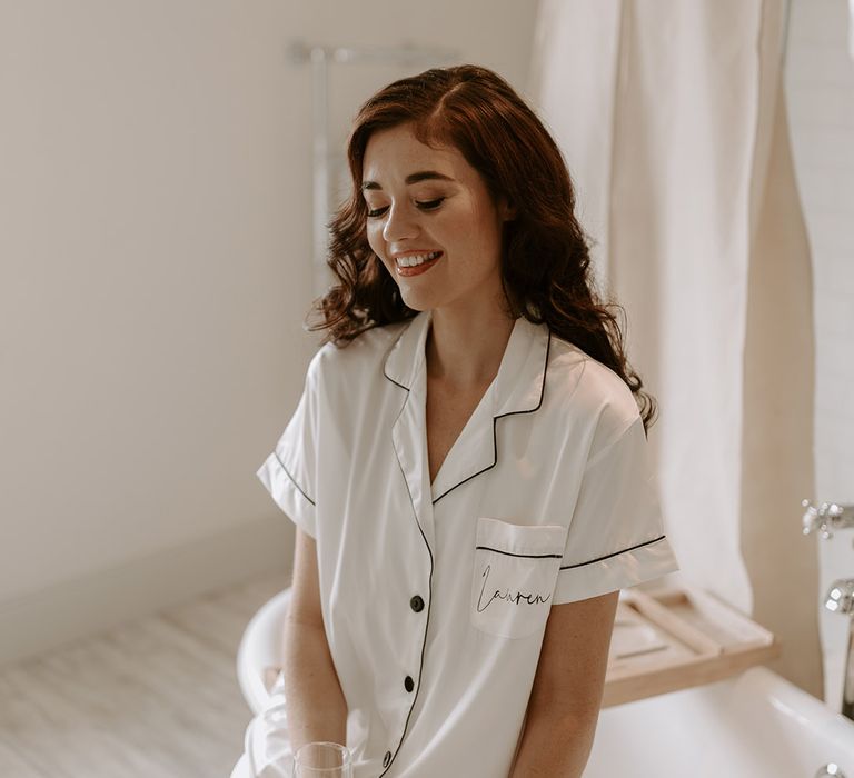 Bride sits on the edge of the bathtub in white satin personalised pyjamas with curled red hair holding glass of champagne 