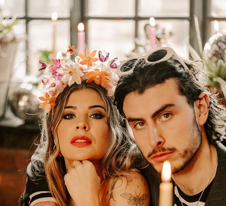 Bride in artistic floral crown and groom in striped black and white long sleeve t shirt with large white sunglasses at colourful grunge glam wedding tablescapae with pink and cream tapered candles 