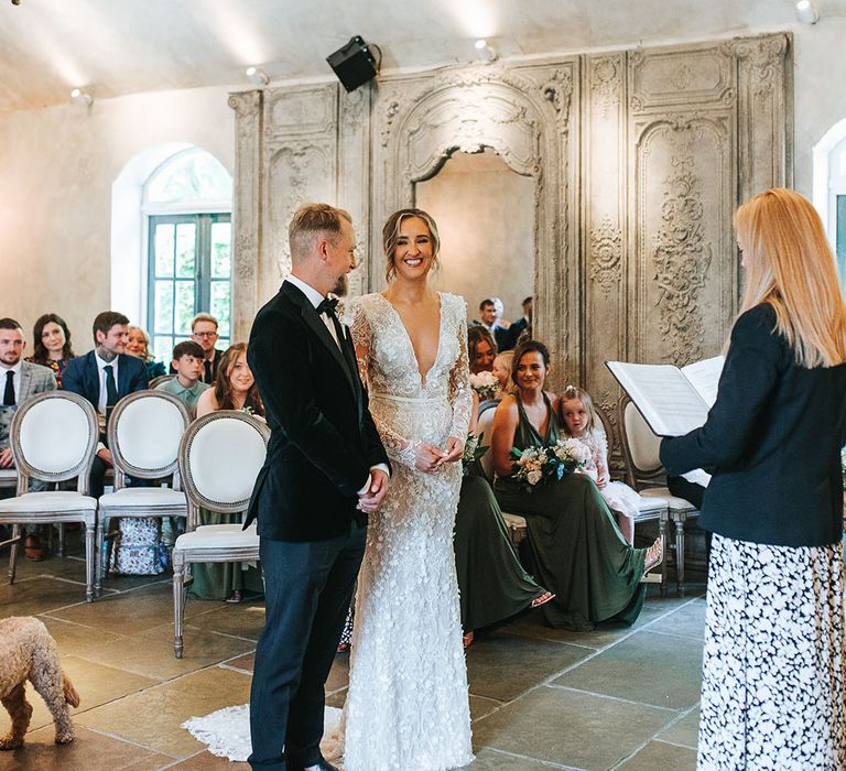 Bride in a 3D lace wedding dress with shoulder pads smiling at the groom as they stand together at the altar with the groom in a black tuxedo 