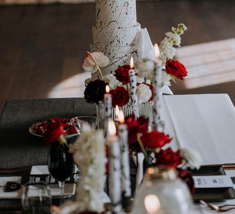 Black red and white wedding decor - red and white roses, carnations and chrysanthemums, black and white tapered candles in clear candlestick holders, small black statues and three tier white wedding cake with black paint splatter icing  