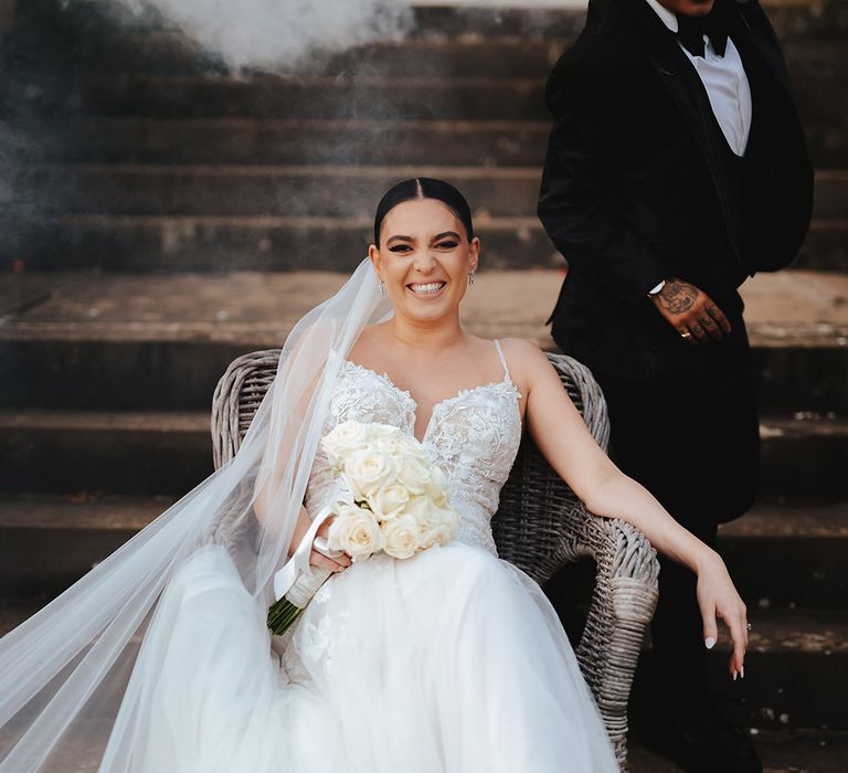 Bride in lace sleeveless v neck Enzoani wedding dress with tulle finish and church length veil sitting on wicker chair during smoke bomb photoshoot at Grittleton House wedding venue 