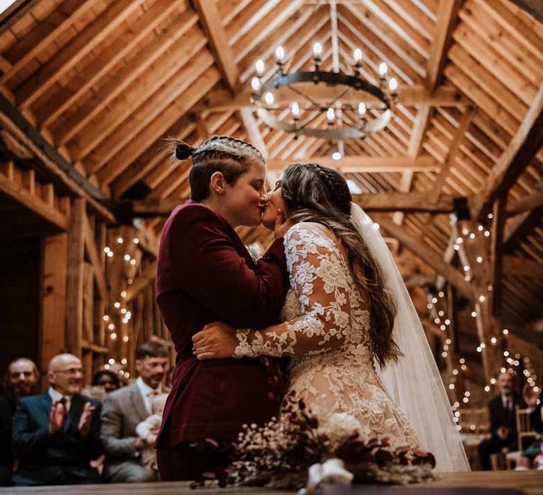 Bride in lace illusion sleeve wedding dress with overlay doing first kiss with bride in maroon bridal suit at LGBTQ+ wedding at Bassmead Manor Barns wedding venue 