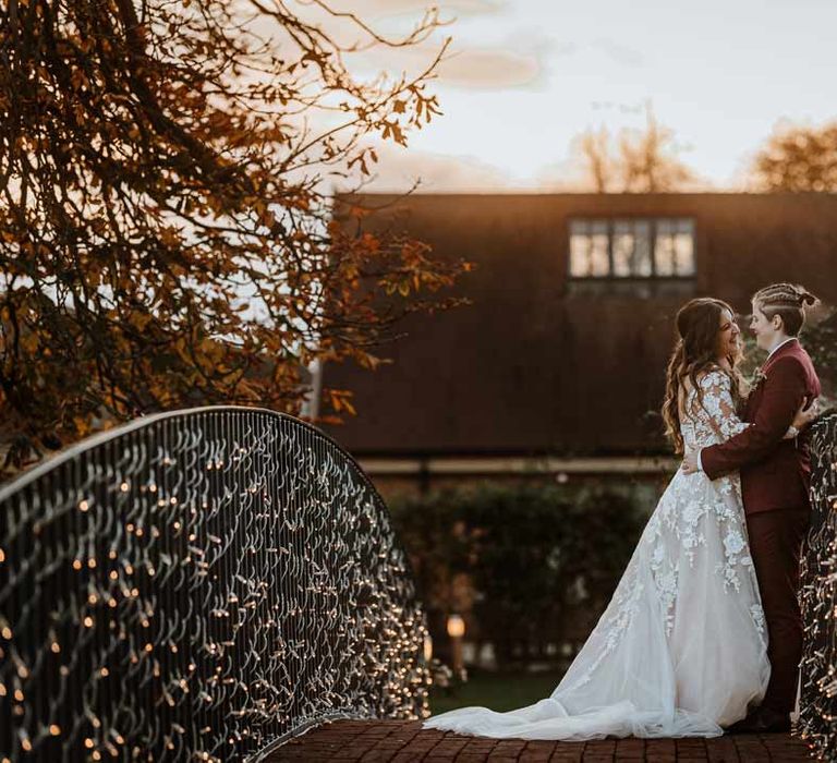 Bride in long sleeve lace illusion sleeve wedding dress with overlay standing on a bridge at Bassmead Manor Barns with bride in maroon bridal suit with grey waistcoat, dusky pink tie and autumnal dried flower boutonniere 