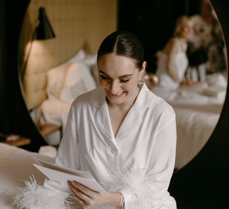 Bride in white bridal pyjamas with feather cuffs reads a letter on the morning of the wedding day 