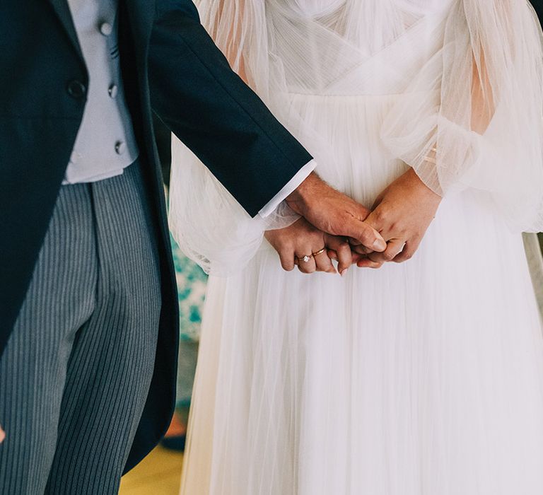 Groom in pinstriped trousers and morning suit holding hands with bride in tulle wedding gown at barn wedding