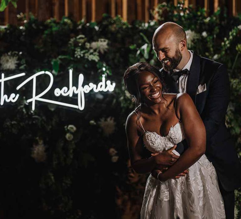 Bride in sweetheart neckline lace wedding dress standing and laughing with groom in classic black three piece grooms suit with bow tie and white rose and foliage boutonniere in church in front of foliage wall and large neon wedding sign 