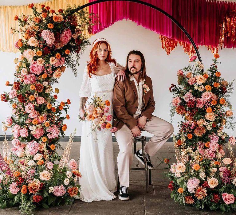 Bride in three quarter length puff sheer sleeve wedding dress with delicate polka dot overlay and square neck and white 3D applique polka dot headband posing with groom in brown blazer, white shirt, grey suit trousers and vans shoes underneath circular floral arch with vibrant flowers and foliage attached and pink, red, yellow and metallic wedding streamers behind