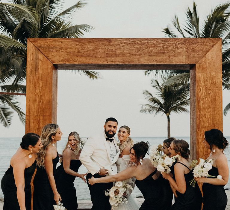 bride in a strapless tulle wedding dress, groom in a white tuxedo jacket, and bridal party in strapless, satin, black bridesmaid dresses standing in front of a wooden frame on the beach 