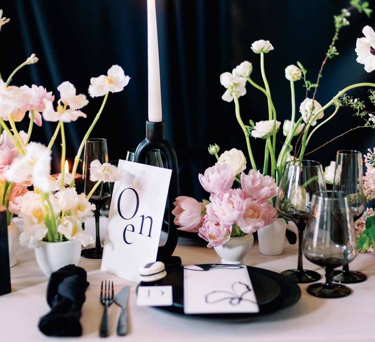 Black fabric backdrop with light pink wedding tablecloth, white poppies, light pink Persian buttercups, baby's-breath and foliage centrepieces, black artistic candlestick holders, white tapered candles, black crockery and monochrome wedding stationery 