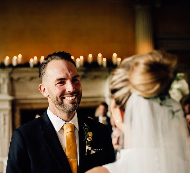 Groom in navy suit with yellow tie and yellow buttonhole smiling at the bride in a plunging wedding dress 