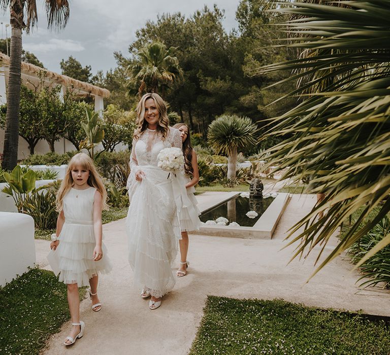 boho bride in delicate lace wedding dress holding a white rose bouquet walking through pure house ibiza wedding venue with her flower girls