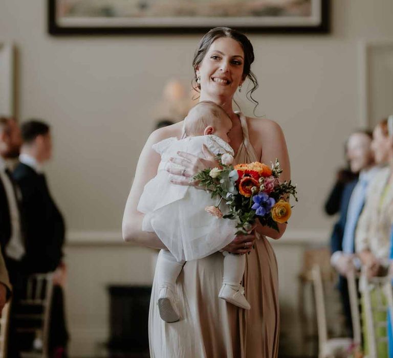 Bridesmaid in multiway off-white bridesmaid dress holding baby walking down the aisle at Elmore Court Gloucestershire