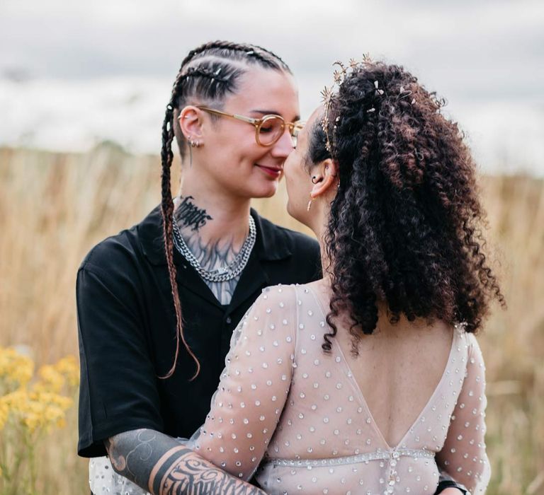 Bride in silver chains, glasses, black shirt and black trousers hugging bride in long sleeve v-neck sheer polka dot overlay sparkly wedding dress in the fields of their at-home Viking themed wedding