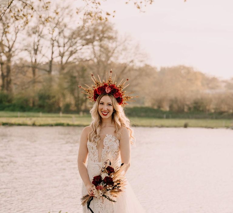 Bride in off shoulder lace wedding dress with corset style top and red rose and gold halo bridal crown holding red rose, pampas grass and dried flower bridal bouquet on the grounds of Southlands Barn