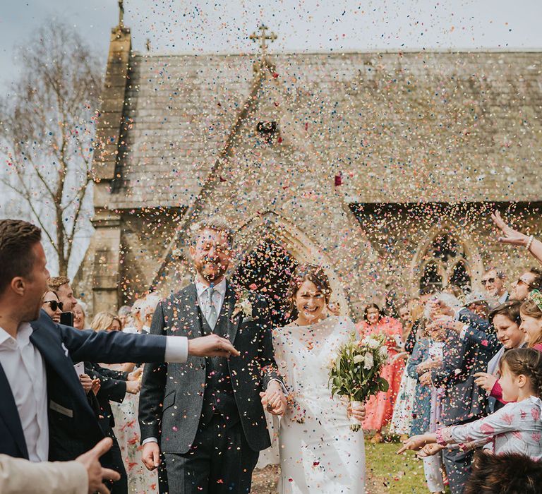 The bride and groom walk out of the church to bright and colourful wedding confetti 